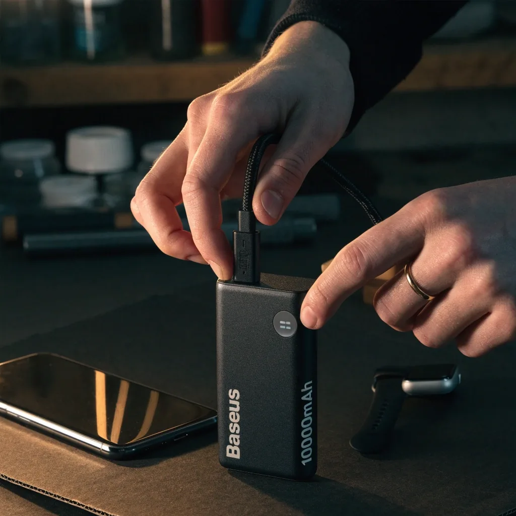 Power bank sobre una mesa de cartón negro con cable USB-C enroscado y un teléfono boca abajo, luz cálida lateral